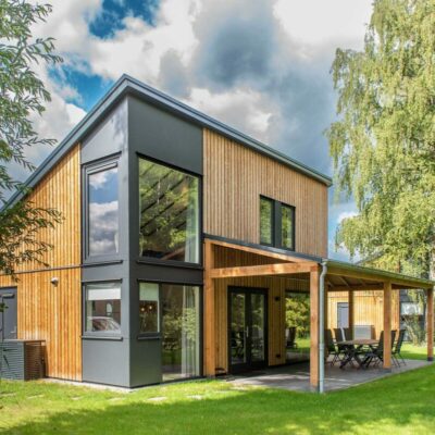 Een modern vakantiehuisje met sterk schuin aflopend dak, op een groen grasveld met groene bomen er omheen.