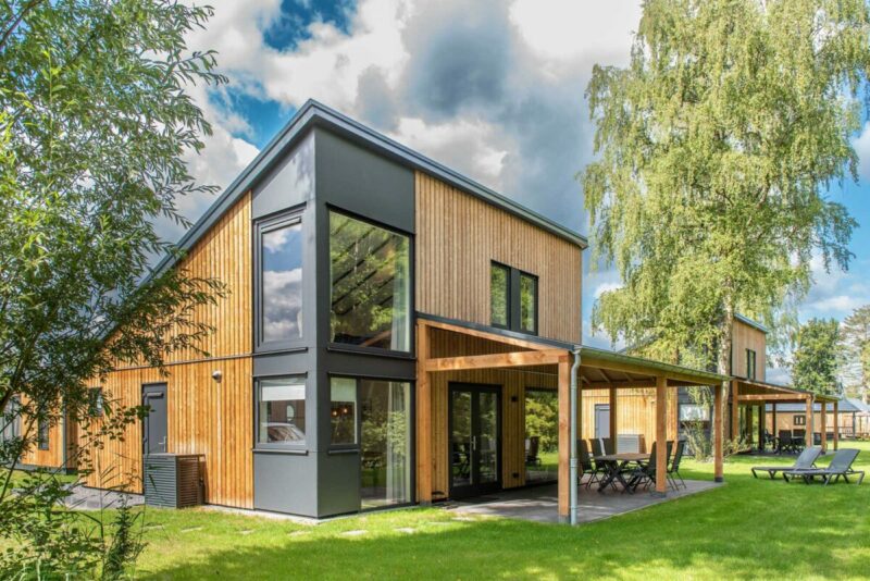 Een modern vakantiehuisje met sterk schuin aflopend dak, op een groen grasveld met groene bomen er omheen.